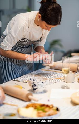 Jeune femme en tablier debout à la table avec divers outils et formant un vase en argile fait main pendant la poterie Banque D'Images