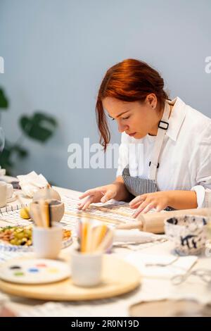 Jeune femme artisanale concentrée dans un tablier avec des cheveux rouges roulante morceau d'argile tout en créant de la poterie dans l'atelier d'artisanat pendant la journée de travail Banque D'Images