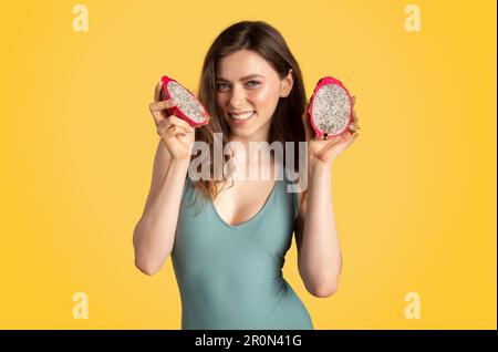 Jolie jeune femme en maillot de bain tenant des tranches de pitaya, appréciant un savoureux fruit juteux de dragon sur fond jaune de studio Banque D'Images