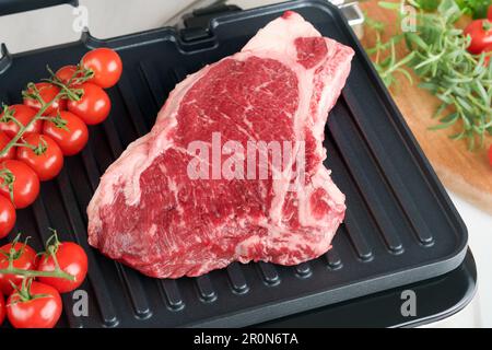 Steaks de première qualité crus sur le gril électrique. Steaks de viande de choix black angus Ribeye sur un grill électrique moderne au romarin, à la tomate cerise et au poivre. Cuisine pro Banque D'Images