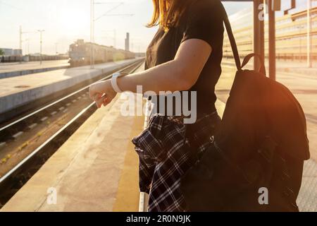 Femme debout à la gare le soir et regardant la montre à la main, train en arrière-plan. Banque D'Images