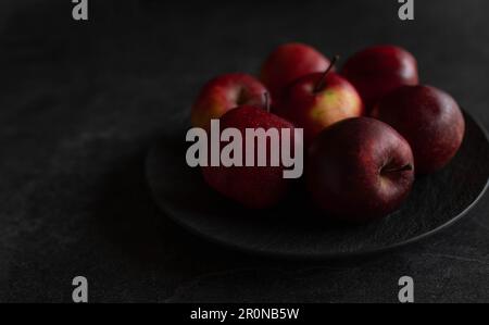Pommes rouges sur une plaque noire isolée sur fond sombre et moody avec espace de copie. Faible luminosité Banque D'Images