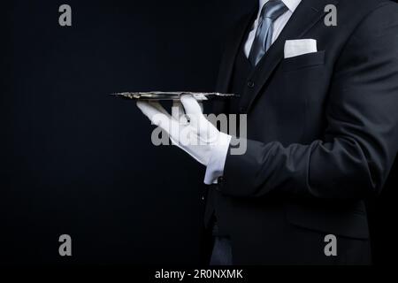 Portrait d'un élégant maître d'hôtel ou d'un serveur en costume sombre et gants blancs tenant un plateau de service argenté. Banque D'Images