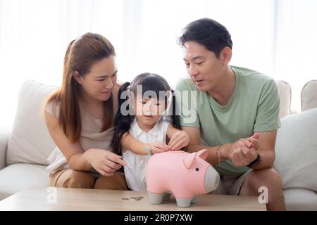 Père de famille asiatique heureux et mère et fille assis sur canapé planification de financement avec des économies tout en déposant des pièces dans la chambre, maman et papa Banque D'Images