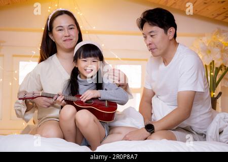 Joyeux père de famille asiatique et mère assis enseignant jouer ukulele guitare avec fille ensemble sur le lit dans la chambre à la maison, maman et papa se détendre avec lea Banque D'Images