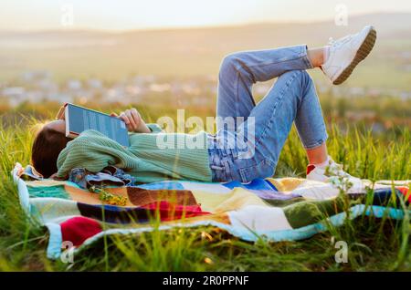 Adolescente avec un livre couvrant la face, se reposant sur une colline au coucher du soleil Banque D'Images