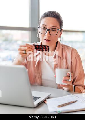 Femme regarde les pilules de médicaments tout en travaillant avec un ordinateur portable. Problèmes de santé mentale, épuisement émotionnel ou maux de tête. Bureau moderne au centre de travail. BT Banque D'Images