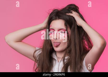 Portrait d'une jeune femme adulte ludique avec les yeux fermés, la langue qui dépasse et les mains relevées à la tête. Beauté jeune femme adulte avec maquillage de scène rose Banque D'Images