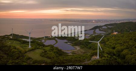 Éoliennes et panneaux solaires avec des lumières de ville lointaine au coucher du soleil Banque D'Images