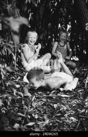 Trois enfants jouent dans une cabane qu'ils ont eux-mêmes construite à partir de feuilles et de brindilles. Photographie en noir et blanc Banque D'Images