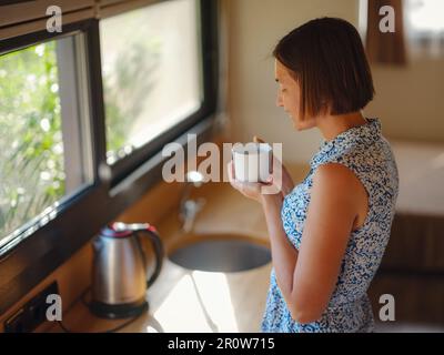 Jeune femme appréciant une tasse de café le matin, dans sa cuisine. Minimalisme. Entrée en marche. Vivre seul. Charmante caravane Banque D'Images