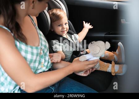 Femme avec un jouet tricoté près de sa fille dans un siège d'enfant à l'intérieur de la voiture Banque D'Images