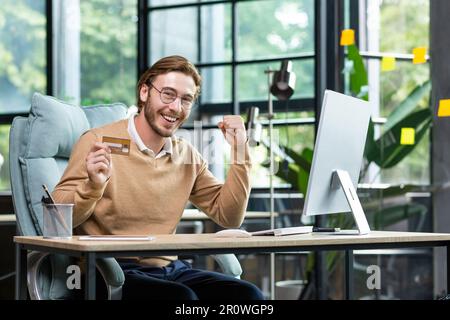 Un jeune homme utilise avec succès une carte de crédit. Il s'assoit heureux dans le bureau à l'ordinateur, tient une carte, regarde la caméra, montre un geste oui, fête. Banque D'Images