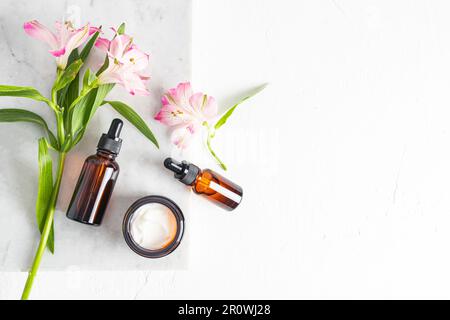 Deux flacons cosmétiques avec compte-gouttes avec produit naturel de soin du visage et un pot ouvert de crème sur un podium en marbre avec fleurs printanières. Vue de dessus Banque D'Images