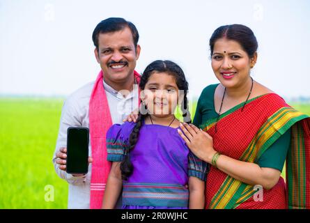 Joyeux village souriant couple avec un enfant montrant un téléphone mobile à écran vert en pointant le doigt tout en regardant l'appareil photo sur les terres agricoles - concept de publicité Banque D'Images