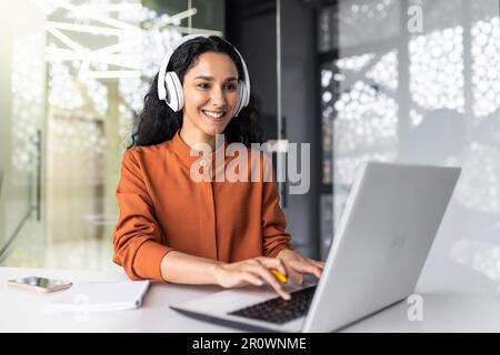 Jeune femme hispanique à succès travaillant au bureau avec des écouteurs, une femme programmant un logiciel de codage sur un ordinateur portable souriant, écoutant des livres audio et des podcasts en ligne, femme d'affaires au bureau. Banque D'Images