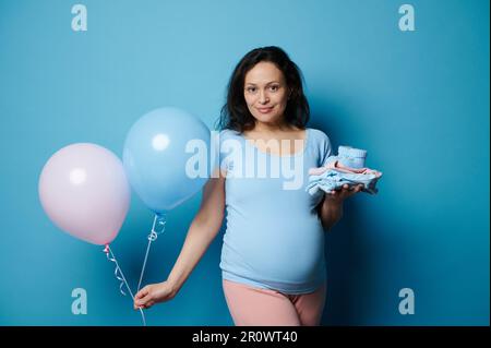 Belle femme enceinte brune posant avec des vêtements nouveau-nés et des ballons d'air bleu rose, isolés sur fond bleu Banque D'Images