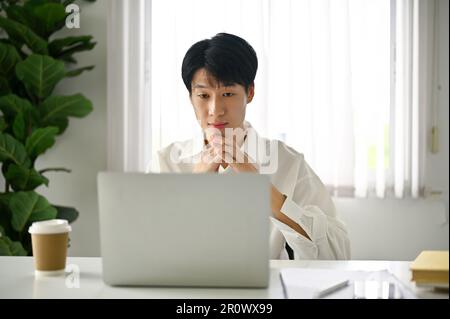 Un jeune homme d'affaires ou un employé de bureau asiatique intelligent et ciblé regardant son écran d'ordinateur portable, planifiant son projet, travaillant dans son bureau. Banque D'Images