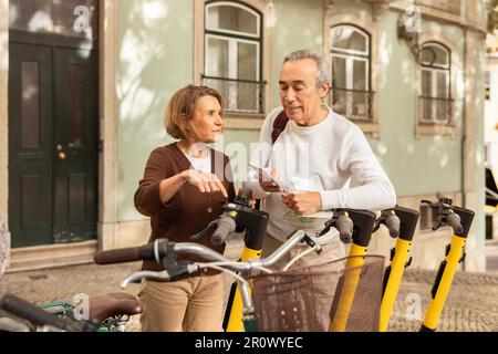 Les voyageurs âgés se coupent à l'aide d'un smartphone louant des scooters électriques en plein air Banque D'Images