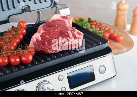 Steaks de première qualité crus sur le gril électrique. Steaks de viande de choix black angus Ribeye sur un grill électrique moderne au romarin, à la tomate cerise et au poivre. Cuisine pro Banque D'Images