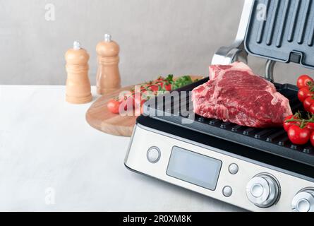 Steaks de première qualité crus sur le gril électrique. Steaks de viande de choix black angus Ribeye sur un grill électrique moderne au romarin, à la tomate cerise et au poivre. Cuisine pro Banque D'Images