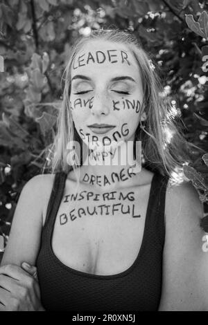 Une jeune femme avec ses yeux fermés et une expression réfléchie avec des mots de motivation écrits sur sa peau, noir et blanc photo Banque D'Images