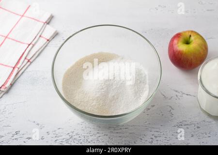 Un bol en verre avec un mélange de farine de blé et de riz, une pomme et un yogourt sur fond bleu clair. Étape de cuisson de délicieuses crêpes faites maison Banque D'Images