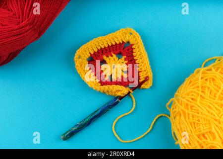 Carré Granny en jaune et rouge étant crocheté, avec des boules de fils en jaune et rouge partiellement visibles dans les coins, le tout sur fond bleu aqua. Banque D'Images