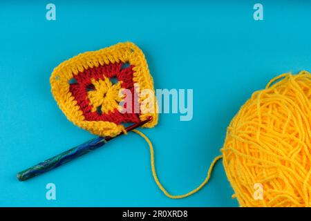 Carré Granny en jaune et rouge étant crocheté, avec une boule de fil en jaune partiellement visible, le tout sur fond bleu aqua. Banque D'Images