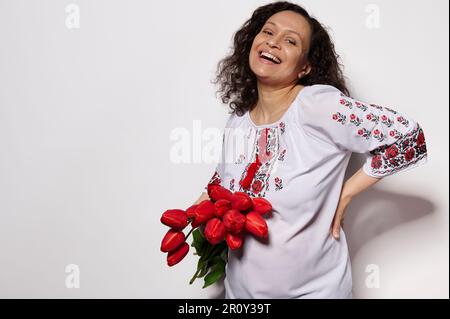 Sourire riant bonne jeune femme enceinte en robe ukrainienne ethnique brodée, avec beau bouquet de tulipes rouges Banque D'Images