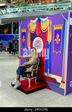 Une jeune femme blonde attirante prenant un selfie tout en étant assise devant un décor célébrant le couronnement du Roi Charles Waterloo Station UK Banque D'Images