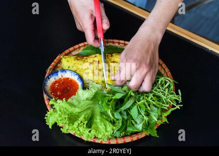 Mains avec des ciseaux coupant crêpes vietnamiennes Banh xeo avec de la nourriture de mer sur fond noir vue de dessus Banque D'Images