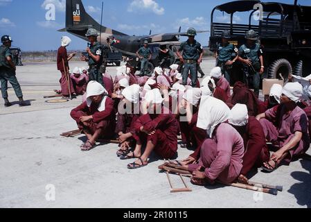 Prisonniers de Viet Cong s'asseoir sur la piste à la base aérienne de Tan Son Nhut, sous le regard vigilant de la police militaire du Sud Vietnam. Les prisonniers ont été amenés à la base aérienne dans le 6X6 camions dans l'arrière-plan et seront transportés par avion à Loc Ninh, Vietnam du Sud sur l'avion de transport C-123 pour l'échange de prisonniers entre les États-Unis et le Vietnam du Sud et Nord Vietnam/Viet Cong militaires. Banque D'Images