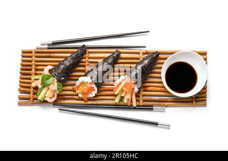 Tapis en bambou avec de savoureux cornets à sushis, sauce soja et baguettes sur fond blanc Banque D'Images