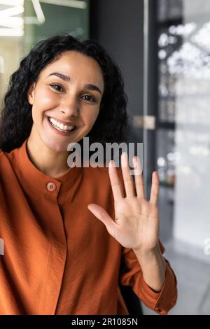 Photo verticale, jeune femme d'affaires hispanique aux cheveux bouclés qui parle lors d'un appel vidéo, agitant la caméra du smartphone, à l'aide d'une application de communication à distance. Banque D'Images