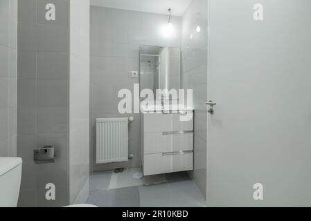 Une armoire simple avec une petite armoire en bois blanc avec des tiroirs sous un miroir sans cadre carré et des carreaux gris brillant Banque D'Images