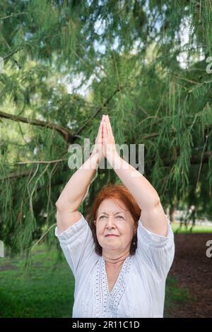 Femme hispanique senior vêtue de vêtements blancs pratiquant le yoga à l'extérieur dans un parc. Vrkssasana debout. Concepts : bien-être, vitalité, actif et sain l Banque D'Images