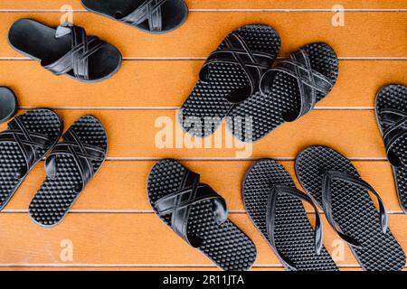 Vue de dessus de plusieurs paires de sandales noires classiques sur fond de bois peint orange sur le patio. De nombreuses paires de tongs en caoutchouc à côté de la piscine ouverte Banque D'Images