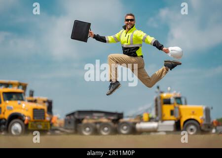 Saut excités d'un constructeur d'âge moyen sur le chantier. Travailleur de construction de constructeur excité dans un casque de sécurité sautant devant les camions. Excitée Banque D'Images