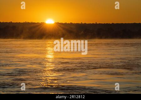 Lever de soleil au-dessus de la rivière Zambèze, la surface de l'eau brille dans des couleurs orange vives, la brume, le brouillard s'élève de l'eau. Zambèze River, Zimbabwe, Afrique Banque D'Images