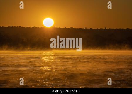 Lever de soleil au-dessus de la rivière Zambèze, la surface de l'eau brille dans des couleurs orange vives, la brume, le brouillard s'élève de l'eau. Zambèze River, Zimbabwe, Afrique Banque D'Images