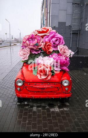 Voiture rétro avec de grandes fleurs près du centre commercial de la ville. Une vieille voiture rouge par temps nuageux. Banque D'Images