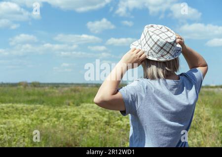 Une femme dans un chapeau et un T-shirt bleu dans un champ, ciel bleu avec des nuages, vue arrière. Heure d'été. Vacances au village. Temps de déplacement Banque D'Images