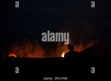 Une figure masculine est silhouetée la nuit par les flammes d'un feu ouvert sur une plage à côté des dunes de reedy Banque D'Images