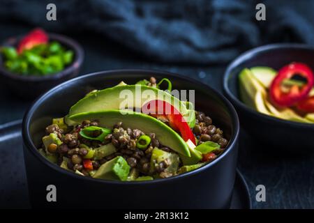Salade de lentilles avec avocat et poivron rouge dans un bol noir sur fond noir. Nourriture végétarienne et végétalienne. Banque D'Images