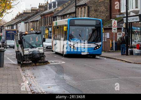 Un nettoyeur de route de Hako Citymaster balaie la gouttière de Mill Road, Cambridge, Royaume-Uni, alors qu'un autobus de Stagecoach passe devant. Banque D'Images