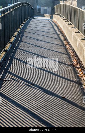 Une passerelle au-dessus de la rivière Derwent à Belper, dans le Derbyshire, en Angleterre, avec des ombres strippées projetées sur la passerelle depuis le soleil qui brille à travers la clôture Banque D'Images