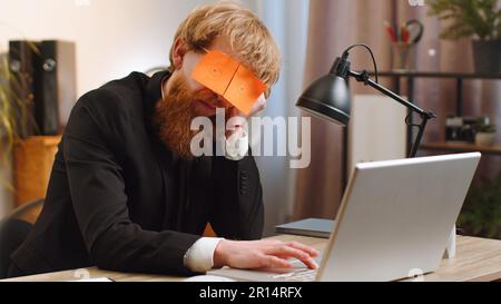 Les gens d'affaires trichent pour dormir. Un jeune homme d'affaires fatigué et inefficace qui dormait sur un ordinateur portable avec des autocollants pour les yeux sur le visage du bureau. Un homme indépendant à la gestion paresseuse barbu Banque D'Images