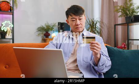 Homme asiatique utilisant la carte de crédit et ordinateur portable tout en transférant de l'argent, achats en ligne, commander la livraison de nourriture à l'appartement à la maison. Homme adulte dans la salle de séjour de jour assis sur un canapé à l'intérieur Banque D'Images