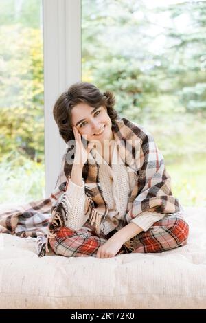 Portrait seule de la jeune femme caucasienne assis près de la fenêtre des appartements dans un chandail blanc et un écossais rouge, passe du temps le matin sur la véranda dans une maison de campagne Banque D'Images